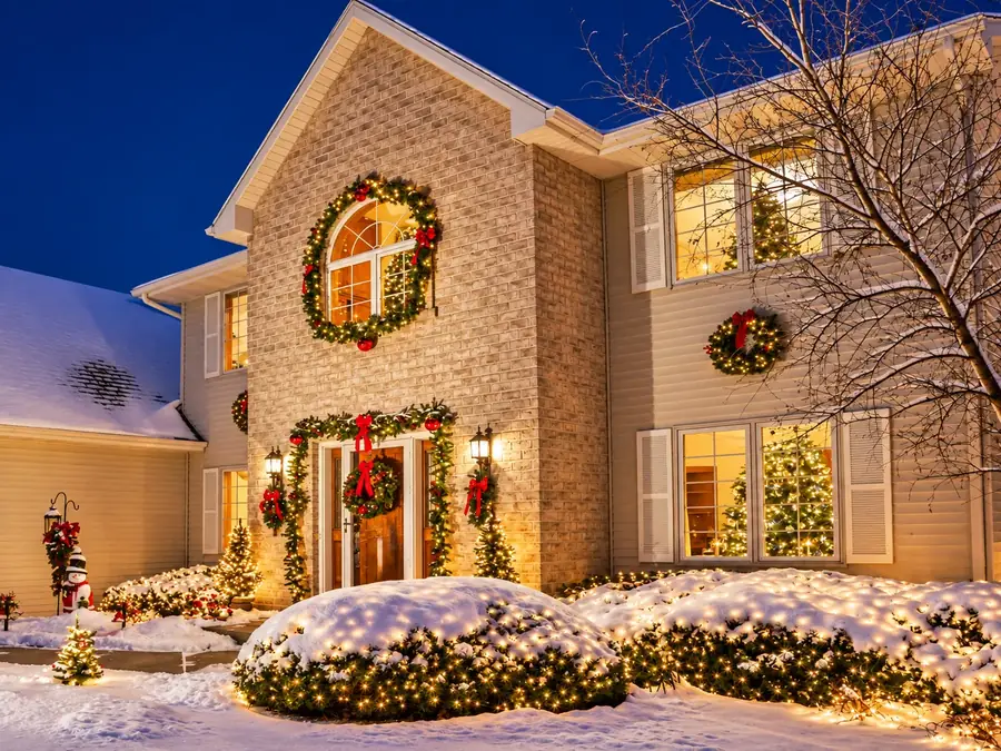 Brick home with lit wreaths, door garland, and net-lit bushes in snow